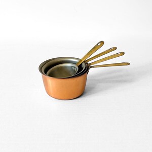May include: A set of four nested measuring cups with copper-toned exterior bodies and metallic handles. Each cup features a round bowl with a silver-toned interior and a long, flat handle with a small circular hole at the end for hanging storage.