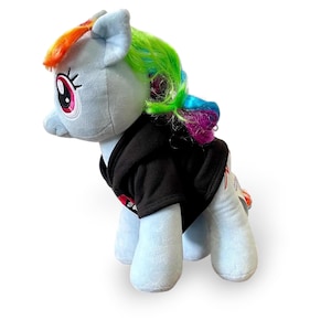 May include: A light blue plush pony with a rainbow mane and tail, wearing a black hoodie. The pony has large pink and black eyes. The plush toy is standing on a white surface.