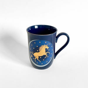 May include: A dark blue ceramic mug featuring a gold unicorn design. The unicorn is set against a blue oval with gold stars. The mug has a gold rim and a curved handle.