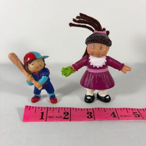 May include: Two Cabbage Patch Kids figurines. One is a baseball player in a blue and red uniform holding a bat. The other is a girl in a purple dress with brown yarn hair and a green glove. A pink measuring tape is in the foreground.