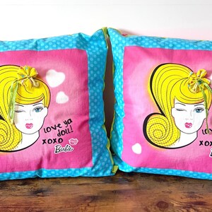 May include: Two square throw pillows featuring a graphic of a blonde character with a high ponytail, winking, and the text "love ya doll! xoxo Barbie". The pillows have a pink center panel with white hearts and a blue border with white polka dots.
