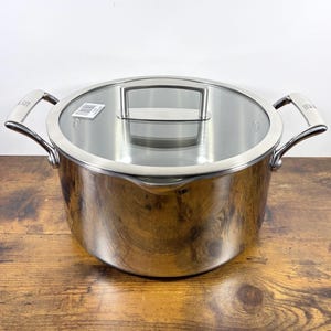 May include: A stainless steel cooking pot with a glass lid. The pot has two curved handles and a clear glass lid with a stainless steel handle. The pot sits on a wooden surface.