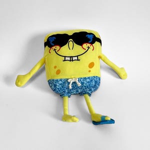 May include: A yellow plush toy shaped like a rectangular character wearing black sunglasses with blue palm tree accents, a wide smiling mouth, and blue patterned swim trunks with a white drawstring. The character has thin yellow arms and legs.
