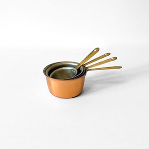May include: A set of four nested measuring cups with copper-toned exterior bodies and metallic handles. The cups feature a rounded shape and are stacked together, showcasing the varying lengths of the attached handles extending outward.