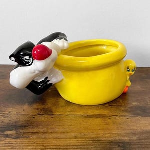 May include: A yellow ceramic planter or bowl featuring a sculpted black and white cat figure with a red nose on one side and a small yellow bird figure with orange feet on the opposite side, both attached to the exterior of the round vessel.
