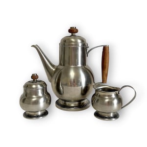 May include: A vintage silver-toned coffee set comprising a teapot, creamer, and sugar bowl. The teapot features a wooden handle and lid finial. The creamer and sugar bowl also have wooden accents. The set is set against a white backdrop.