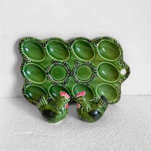 May include: A green ceramic egg tray with 12 egg slots, and two matching chicken-shaped salt and pepper shakers. The tray has a decorative, scalloped edge and a handle. The chickens have pink combs and beaks.