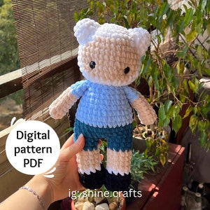 May include: A handmade crochet doll in the likeness of a cartoon character. The doll has a white hat, tan face, blue shirt, teal shorts, and black shoes. The image includes the text "Digital pattern PDF" and the Instagram handle "ig: shine.crafts".