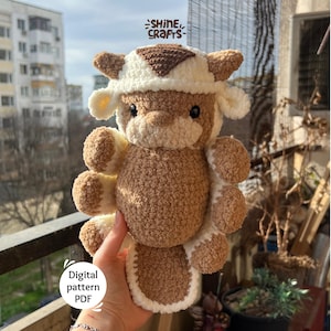May include: A handmade, plush cow toy in shades of brown and white. The toy has a cow-like hat and is held in front of a window. The text "Shine Crafts" is visible. The words "Digital pattern PDF" are also visible.