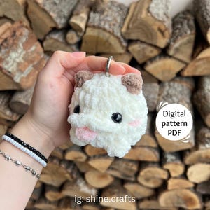 May include: A white crochet plush keychain featuring brown ears, black eyes, and a pink tongue. The plush has a round, fluffy texture and includes a silver metal keyring. Text on the image reads Digital pattern PDF and ig: shine.crafts.