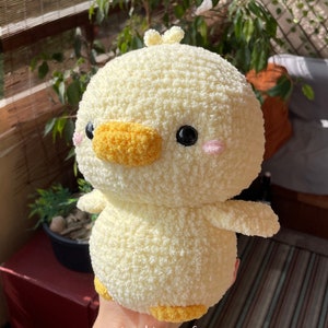 May include: A round, pale yellow crocheted plush duck with a textured, fuzzy yarn finish. It features a bright orange beak, small black bead eyes, and soft pink embroidered cheeks. A small tuft of yarn sits on top. The text ig: shine.crafts is visible.