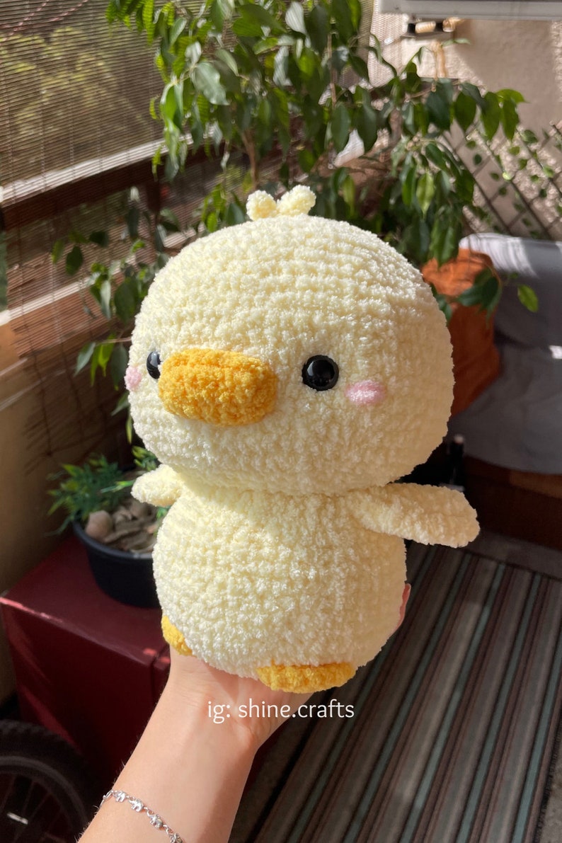 May include: A round, pale yellow crocheted plush duck with a textured, fuzzy yarn finish. It features a bright orange beak, small black bead eyes, and soft pink embroidered cheeks. A small tuft of yarn sits on top. The text ig: shine.crafts is visible.