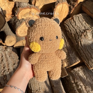 Op de afbeelding: Een bruin pluche speelgoed met een getextureerd, pluizig oppervlak, voorzien van kleine zwarte oren, ronde zwarte ogen, een piepkleine horizontale mond en felgele ronde wangen. De tekst ig:shine.crafts is zichtbaar boven het pluche speelgoed.
