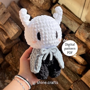 May include: A handmade white crochet doll with black eyes and blush. The doll has a gray cape and black pants. The image includes the text "Digital pattern PDF" and the Instagram handle "ig: shine.crafts".