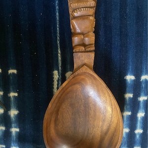 以下が含まれることがあります： ティキの頭が彫刻された木製のサービングスプーン。スプーンは茶色で、滑らかで丸いボウルをしています。