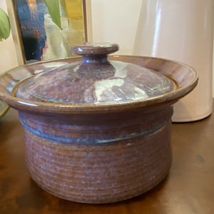 Könnte beinhalten: Eine braune Keramik-Auflaufform mit Deckel. Die Form hat eine blaue und weiße Glasur und eine strukturierte Oberfläche. Der Deckel hat einen Knopf oben.