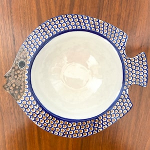 Puede incluir: Un tazón de cerámica blanca con forma de pez con detalles azules y dorados. El tazón tiene un borde festoneado y una superficie texturizada.