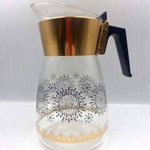Peut inclure: Une cafetière en verre avec une bande dorée autour du haut et une poignée en plastique noir. La cafetière a un motif de flocons de neige noir et or.
