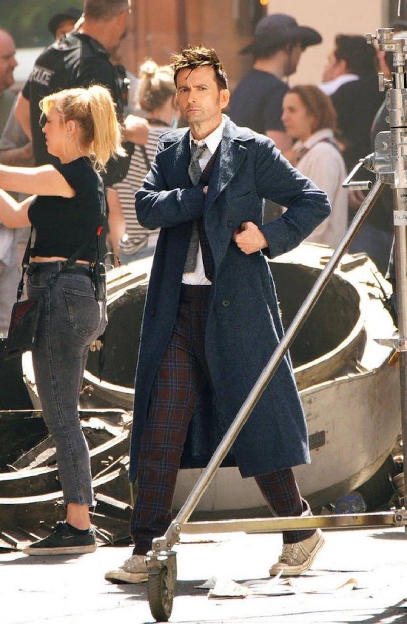 Puede incluir: Un hombre con un abrigo azul sobre una camisa a cuadros y una corbata. Est&aacute; de pie en una acera con un soporte de metal delante de &eacute;l.
