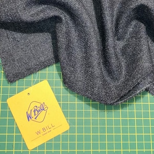Puede incluir: Primer plano de una tela gris oscura con tejido de espiga. La tela est&aacute; doblada y drapeada sobre una estera de corte verde. Una etiqueta amarilla con el texto "W. Bill Since 1848" es visible en la esquina inferior izquierda.
