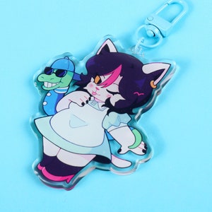 Puede incluir: Un llavero de acrílico colorido con un gato de dibujos animados que lleva un vestido blanco y sostiene un cocodrilo verde con gafas de sol. El gato tiene el pelo rosa y negro y una nariz rosa.