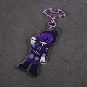 Puede incluir: Llavero acr&iacute;lico morado y negro con un personaje de dibujos animados sosteniendo una calavera. El personaje tiene pelo morado, un gorro negro y un atuendo negro y morado. El llavero tiene un cierre en forma de murci&eacute;lago.