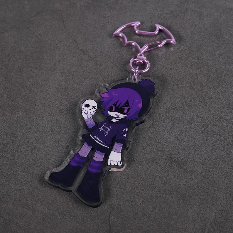 Puede incluir: Llavero acr&iacute;lico morado y negro con un personaje de dibujos animados sosteniendo una calavera. El personaje tiene pelo morado, un gorro negro y un atuendo negro y morado. El llavero tiene un cierre en forma de murci&eacute;lago.