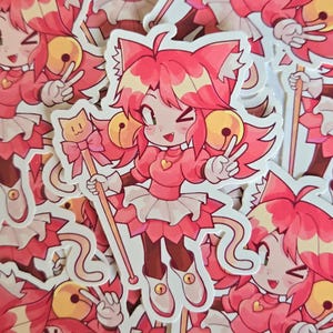 May include: A die-cut vinyl sticker featuring a cartoon character with pink hair, cat ears, and a tail. The character wears a pink outfit with a heart pendant, white gloves, and boots with bells. They hold a staff topped with a cat-shaped emblem.
