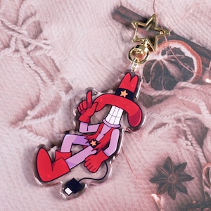 Puede incluir: Un personaje de dibujos animados rojo y morado que lleva un sombrero de vaquero rojo con una estrella dorada. El personaje sostiene una pistola roja y tiene una gran sonrisa. El personaje está unido a un llavero en forma de estrella dorada.