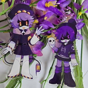 Puede incluir: Dos llaveros acr&iacute;licos con personajes de estilo anime con pelo morado y atuendos negros. Un personaje lleva sombrero y abrigo, el otro sostiene una calavera. Ambos tienen rasgos rob&oacute;ticos, sobre un fondo de flores moradas.