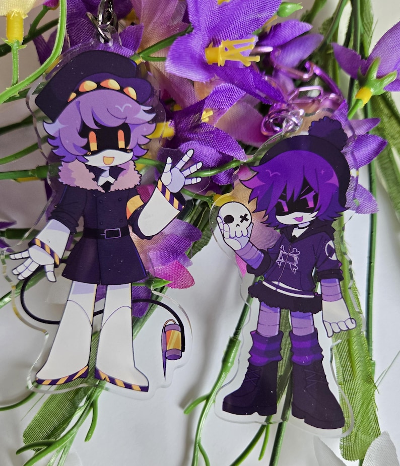Puede incluir: Dos llaveros acr&iacute;licos con personajes de estilo anime con pelo morado y atuendos negros. Un personaje lleva sombrero y abrigo, el otro sostiene una calavera. Ambos tienen rasgos rob&oacute;ticos, sobre un fondo de flores moradas.