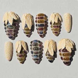 May include: A set of ten press-on nails featuring a variety of designs. Some nails have a textured, cream-colored matte finish, while others display a raised, multi-colored glitter pattern resembling corn kernels topped with cream-colored husks.