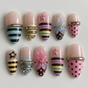 May include: A set of ten almond-shaped press-on nails featuring a nude base with various decorative elements including horizontal stripes in blue, brown, and yellow, pink polka dots, three-dimensional flower appliqués, and small rhinestones.