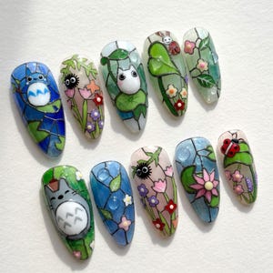 Könnte beinhalten: Ein Set aus zehn mandelförmigen Press-on-Nägeln mit Designs im Buntglas-Stil. Die Motive zeigen Zeichentrickfiguren, Marienkäfer, Rußmännchen und verschiedene Blumenmuster in leuchtenden Blau-, Grün-, Rosa- und Weißtönen.