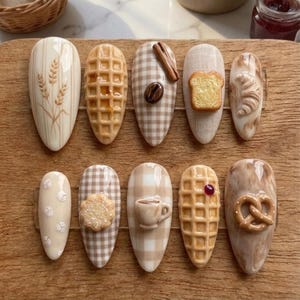 May include: A set of ten artificial nails with various food-themed designs. The nails feature wheat stalks, waffles, coffee beans, toast, a cinnamon stick, a cookie, a cup of coffee, a waffle with a berry, and a pretzel. The color palette includes beige, brown, and tan.