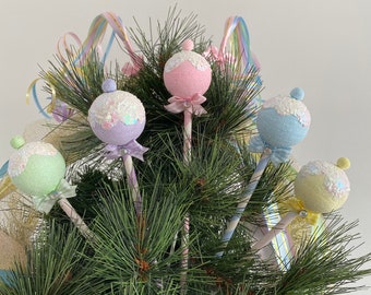 May include: Five pastel-colored ornaments with glitter and bows on a green pine tree. The ornaments are shaped like balls and are attached to sticks.