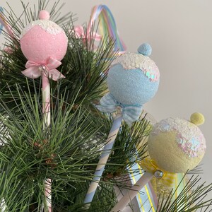 May include: Three pastel-colored Christmas ornaments shaped like lollipops with glitter and ribbon accents. The ornaments are pink, blue, and yellow and are attached to striped paper straws.