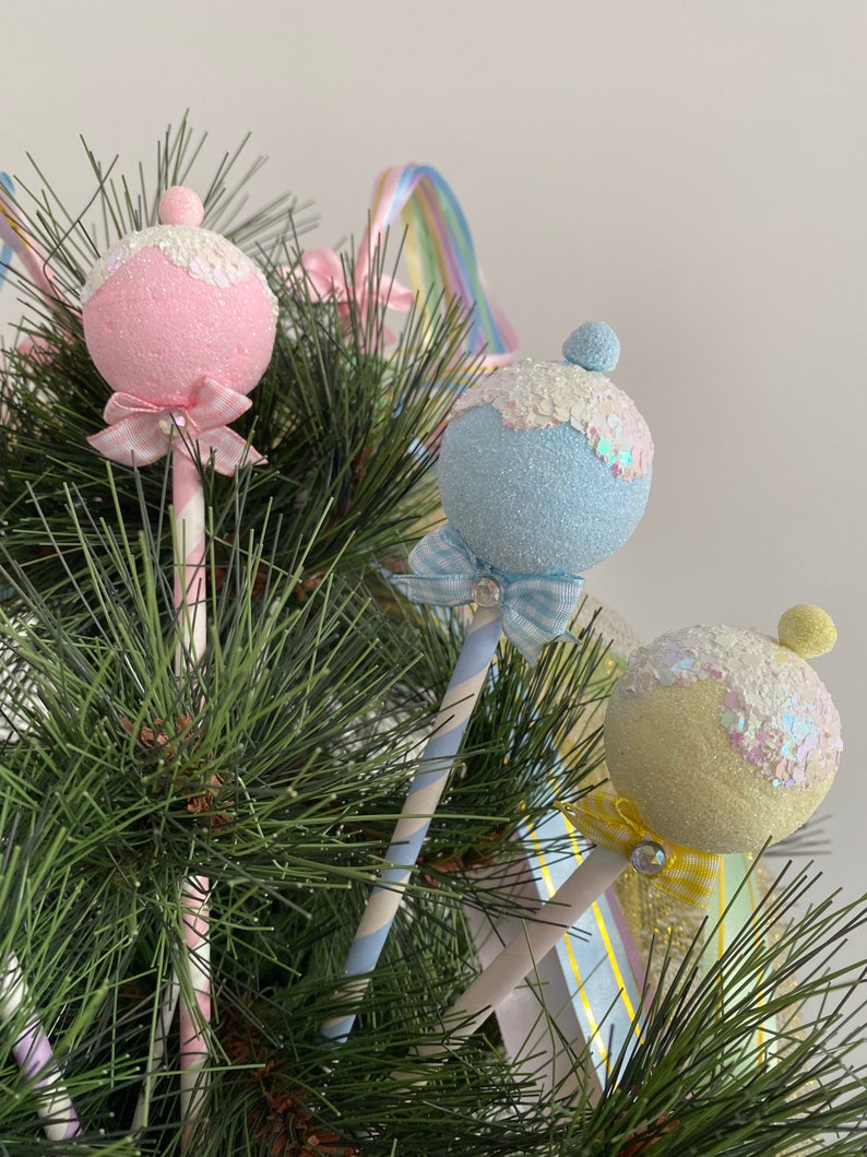 May include: Three pastel-colored Christmas ornaments shaped like lollipops with glitter and ribbon accents. The ornaments are pink, blue, and yellow and are attached to striped paper straws.