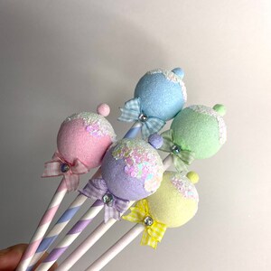 May include: Five pastel-colored, glittery, round ornaments on striped sticks with bows. The ornaments are light blue, pink, lavender, yellow, and green.