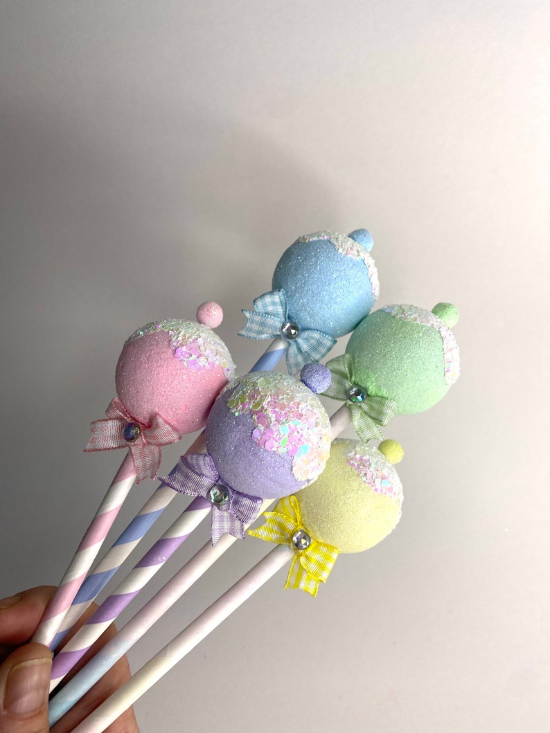 May include: Five pastel-colored, glittery, round ornaments on striped sticks with bows. The ornaments are light blue, pink, lavender, yellow, and green.