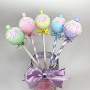 May include: Five pastel-colored, round, decorative ornaments with glitter on top of striped straws in a clear glass jar with a purple bow.
