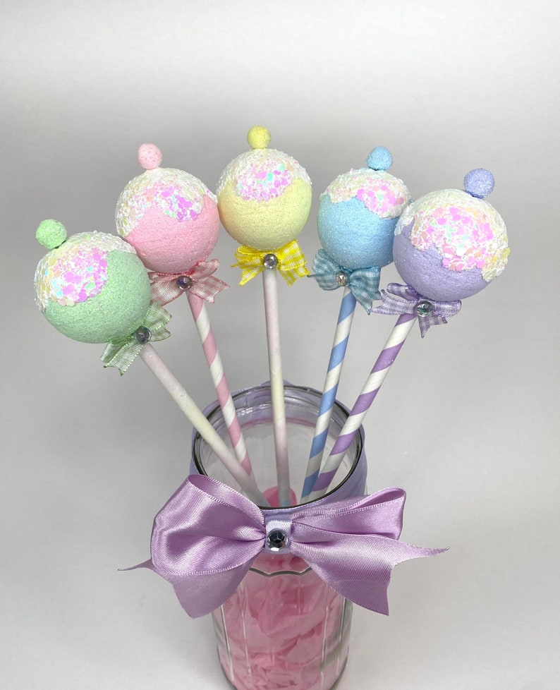 May include: Five pastel-colored, round, decorative ornaments with glitter on top of striped straws in a clear glass jar with a purple bow.