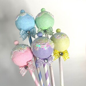 May include: Five pastel-colored, glittery, round ornaments on striped sticks with bows. The ornaments are light blue, green, purple, pink, and yellow.