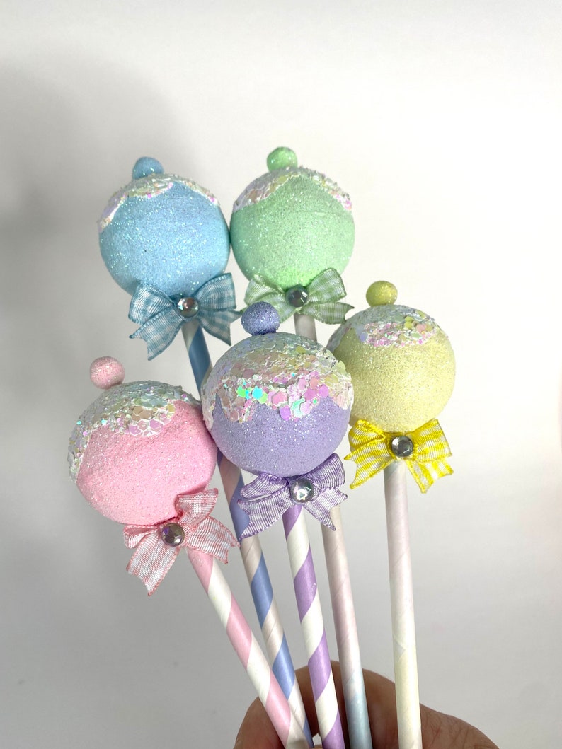 May include: Five pastel-colored, glittery, round ornaments on striped sticks with bows. The ornaments are light blue, green, purple, pink, and yellow.