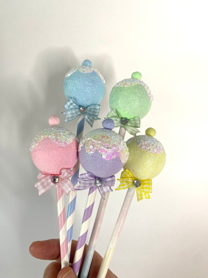 May include: Five pastel-colored, sparkly, round ornaments on sticks with striped paper straws and small bows. The ornaments are light blue, green, pink, purple, and yellow.