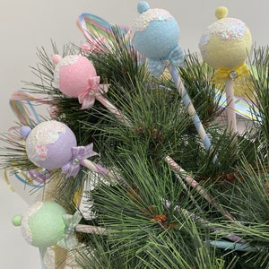 May include: Five pastel-colored ornaments shaped like lollipops with glitter and bows, hanging on a green pine tree branch.