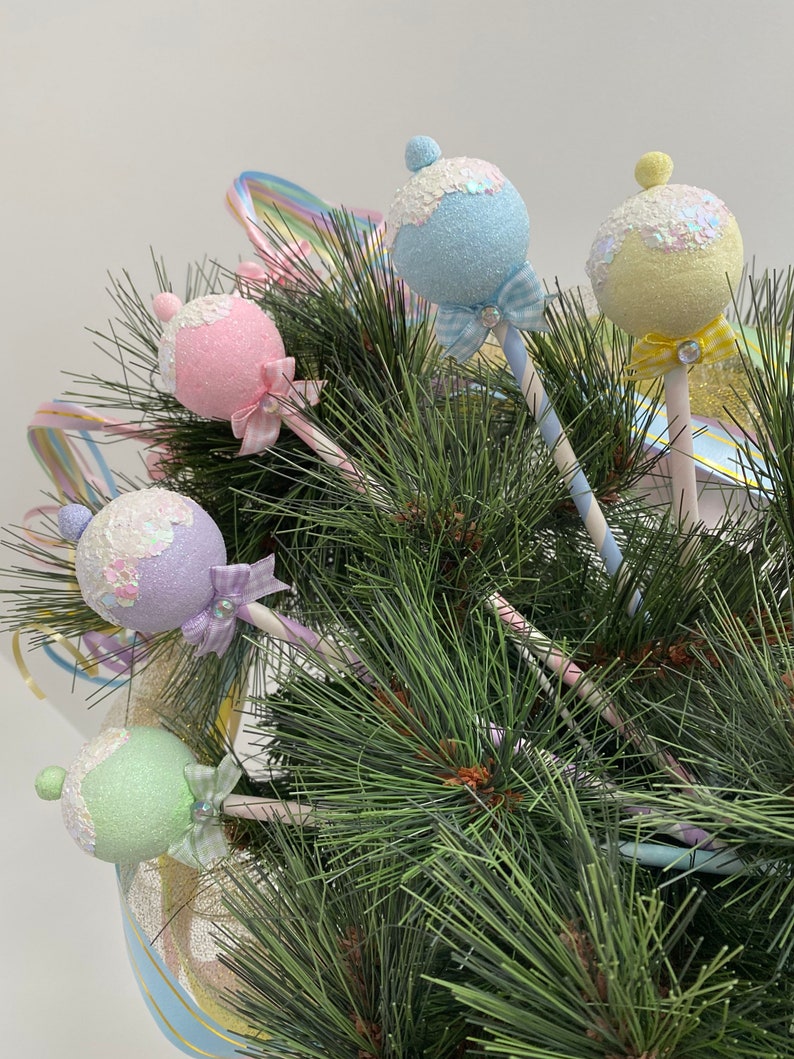 May include: Five pastel-colored ornaments shaped like lollipops with glitter and bows, hanging on a green pine tree branch.