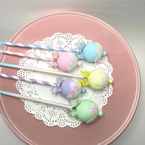 May include: Five pastel-colored cake pops with glitter and bows on sticks. The cake pops are arranged on a white doily on a pink plate.