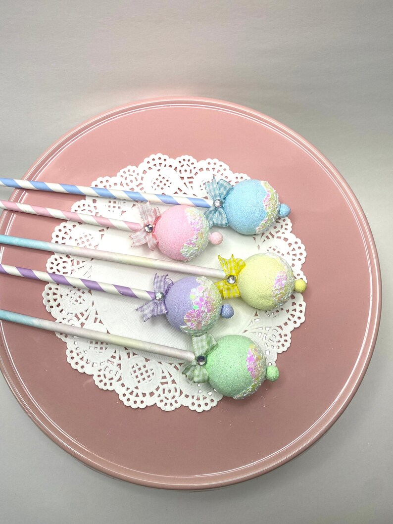 May include: Five pastel-colored cake pops with glitter and bows on sticks. The cake pops are arranged on a white doily on a pink plate.