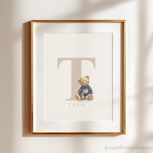 May include: A framed art print with a gold frame. The print features a beige letter "T" and a teddy bear wearing a sweater with an American flag. The name "THEO" is printed below the letter. The background is off-white.